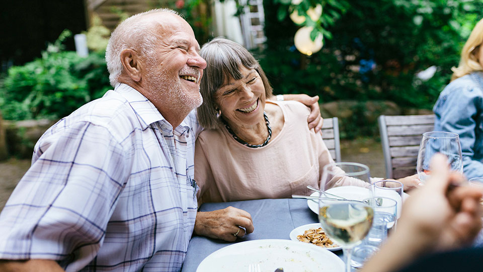 Ein-Mann-legt-einer-Frau-an-einem-Esstisch-im-Garten-den-linken-Arm-und-die-Schulter-und-beide-lachen
