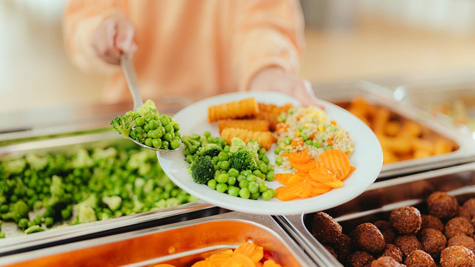 Teller-gefuellt-mit-Kroketten-Couscous-Moehrenscheiben-und-Erbsen-wird-ueber-eine-Speisenausgabe-gehalten