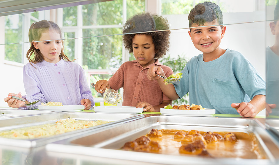 Zwei Mädchen und ein Junge nehmen sich an der Essensausgabe selbstständig Essen