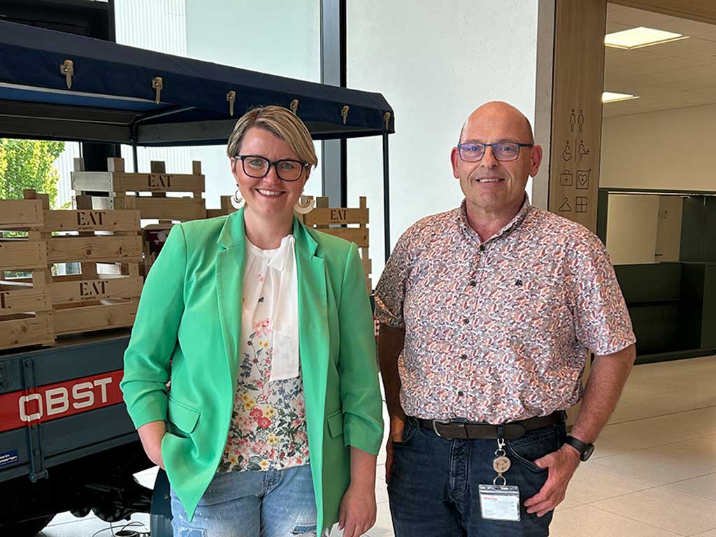 Zwei Personen stehen in einem Innenraum vor einem Wagen mit Holzkisten, die mit ‚EAT‘ beschriftet sind. Die Person links trägt einen grünen Blazer über einem geblümten Shirt und Jeans, die Person rechts ein gemustertes Shirt und dunkle Hose. Beide lächeln und blicken in die Kamera. Im Hintergrund sind ein Bürobereich mit Symbolen an der Wand und große Fenster mit Tageslicht zu sehen.