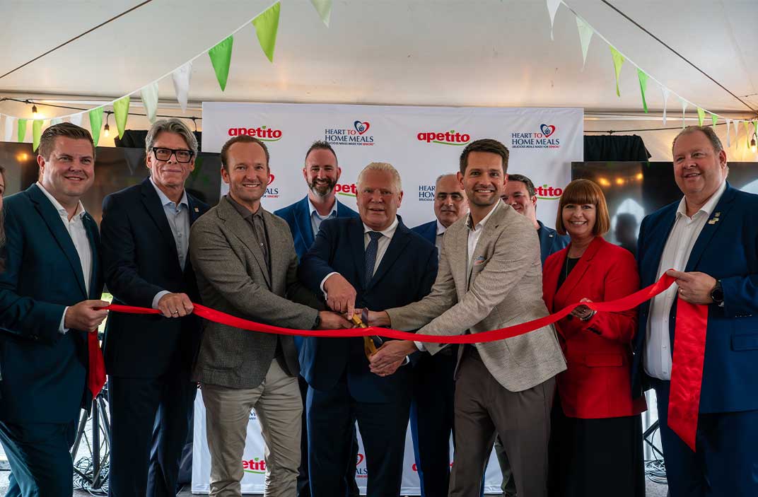 Eine Gruppe von Personen in formeller Kleidung versammelt sich unter einem Zelt zu einer feierlichen Banddurchschneidung. Zwei Personen in der Mitte schneiden ein rotes Band mit goldenen Scheren. Im Hintergrund ist ein Banner mit den Logos von ‚apetito‘ und ‚Heart to Home Meals‘ zu sehen.