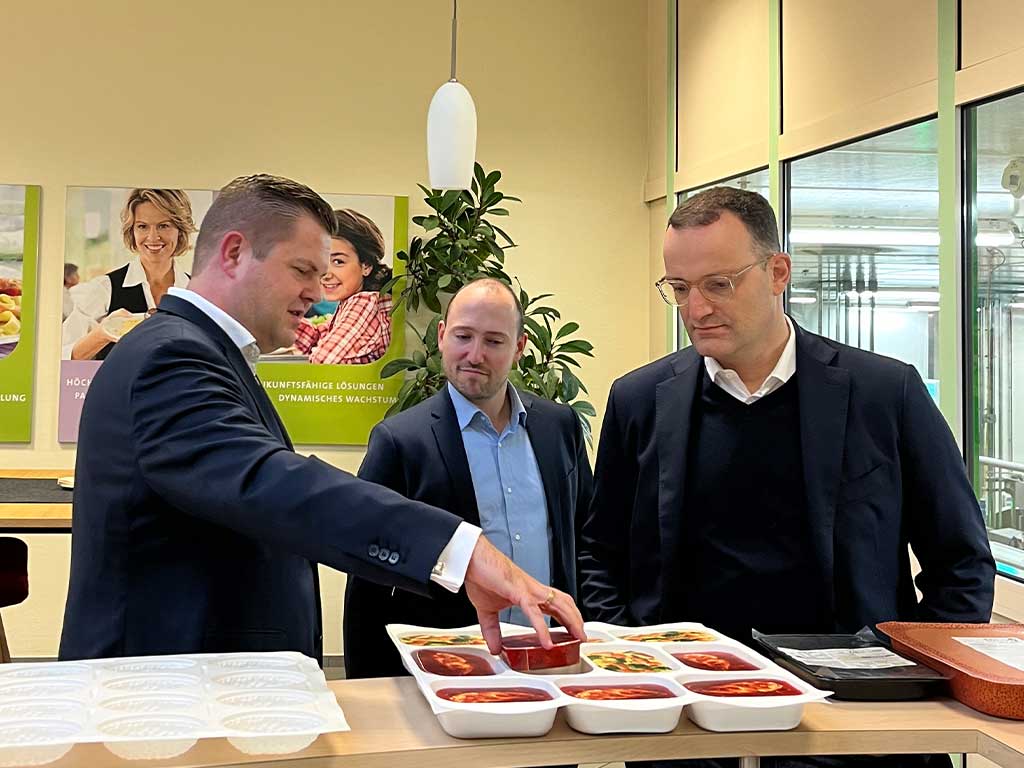 Drei Personen in Anzügen stehen an einem Tisch mit mehreren weißen Tabletts, die verschiedene Gerichte enthalten. Eine Person zeigt auf ein Tablett. Im Hintergrund sind Poster, eine Pflanze und große Fenster sichtbar.