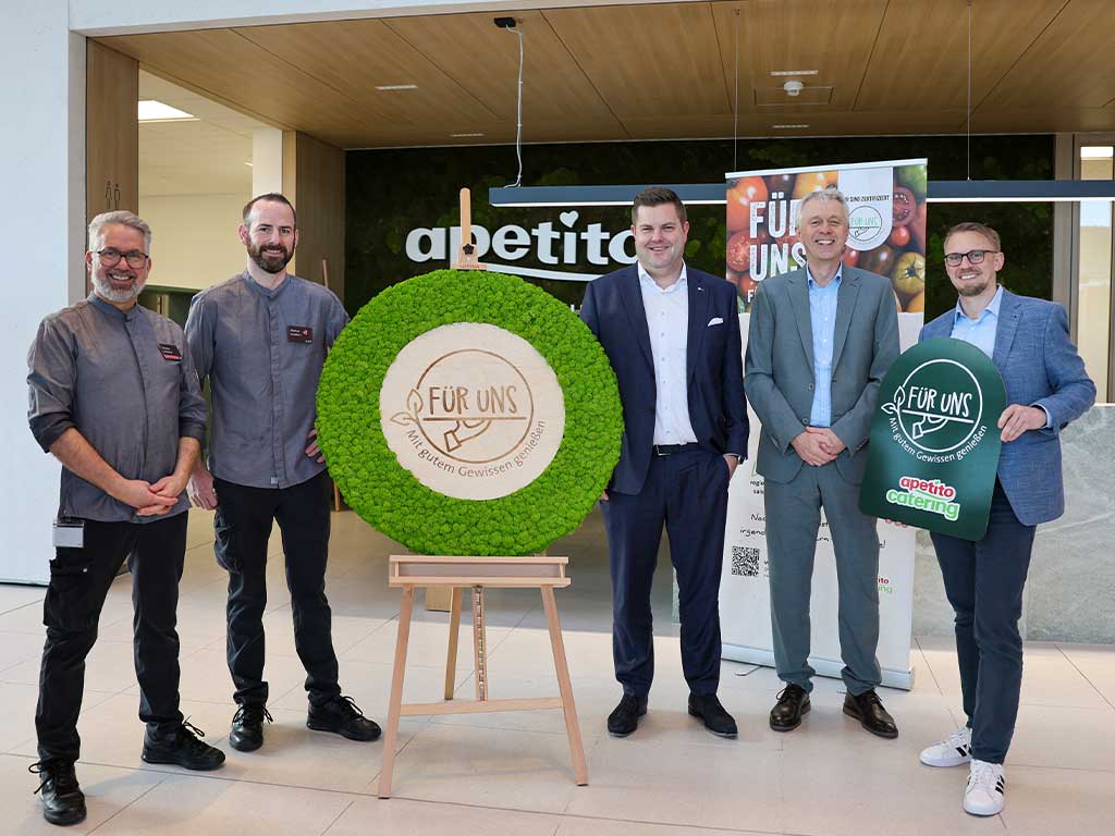 Fünf Personen stehen in einem Eingangsbereich, in der Mitte ein großes „Für Uns“-Logo. Im Hintergrund eine grüne Moos-Wand auf der das weiße apetito-Logo zu erkennen ist.