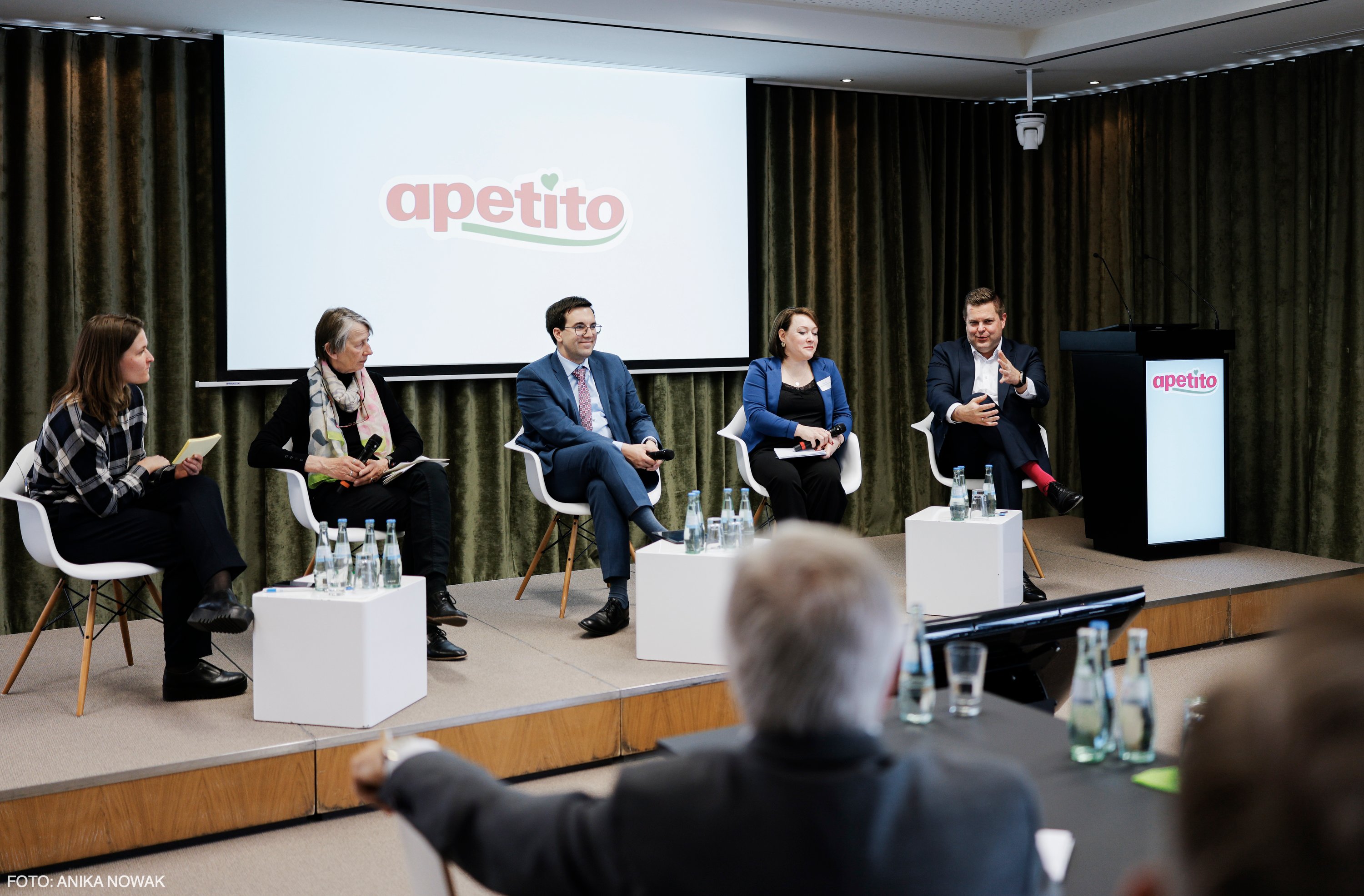 Podiumsdiskussion auf einem apetito Kommunalevent: mehrere Teilnehmende sitzen auf einer Bühne vor einer Leinwand mit apetito Logo, Publikum im Vordergrund.