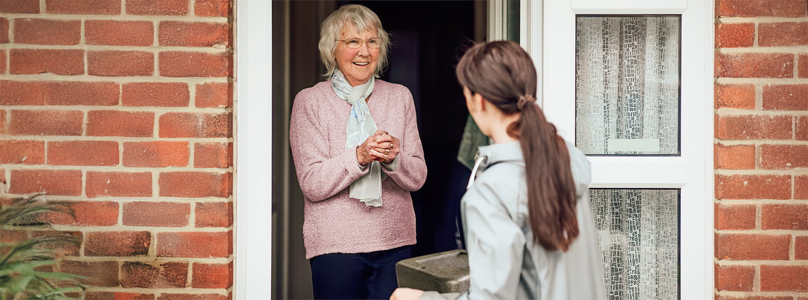 Seniorin steht an der offenen Haustür. Ihr gegenüber steht eine junge Frau mit langen Haaren, die ihr eine Box überreicht